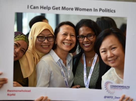 Sign with group of women smiling, reading "I can help get more women in politics".