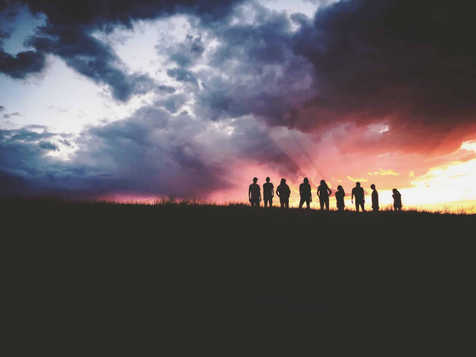 A group of people stand in silhouette beneath dark storm clouds.